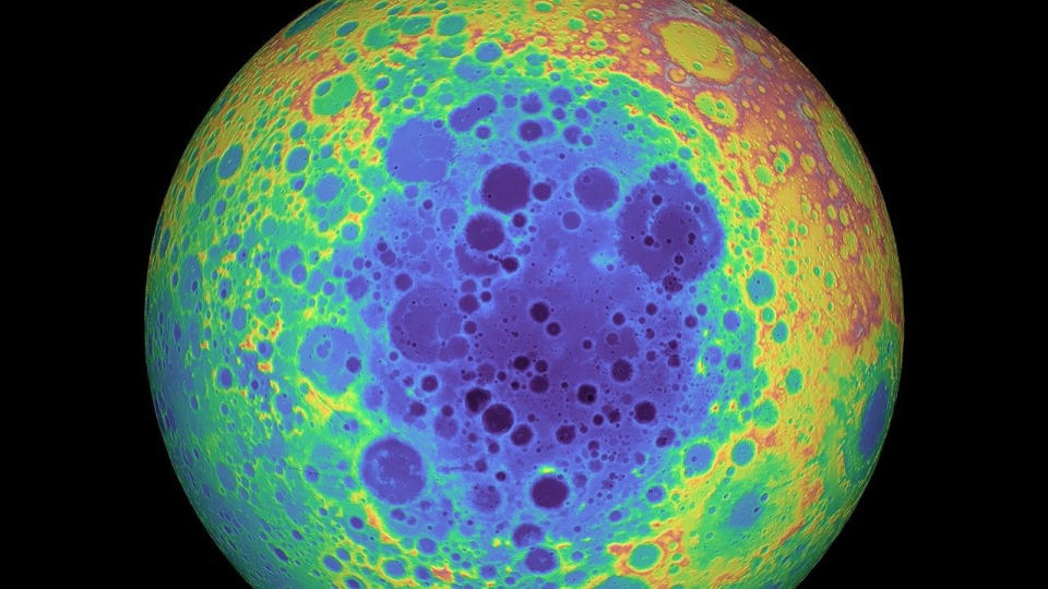  A giant orb takes up the majority of the image, bordered on either sitde by black. Its face is riddled with craters, with colors blotched throughout in vibrant blues, greens, yellows, and reds. 