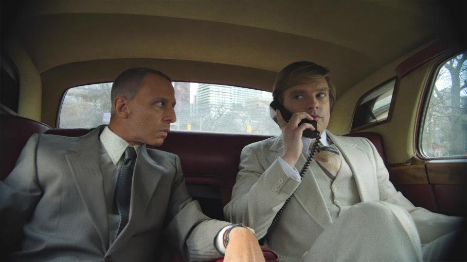 Jeremy Strong looks at Sebastian Stan, who is holding a phone as they both sit in the back of a car, in the movie The Apprentice.