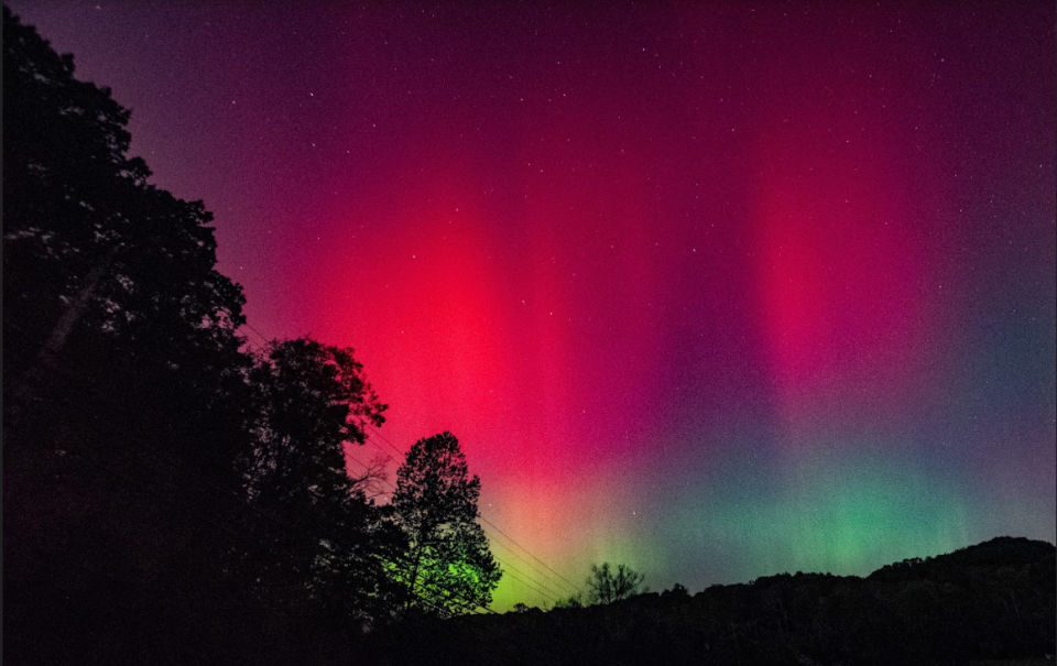  Red and green auroras dance above hills and trees in a dark sky. 