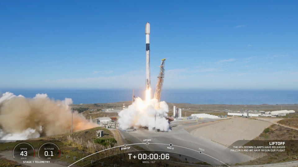  A black and white rocket launches into a clear blue sky from a seaside pad. 