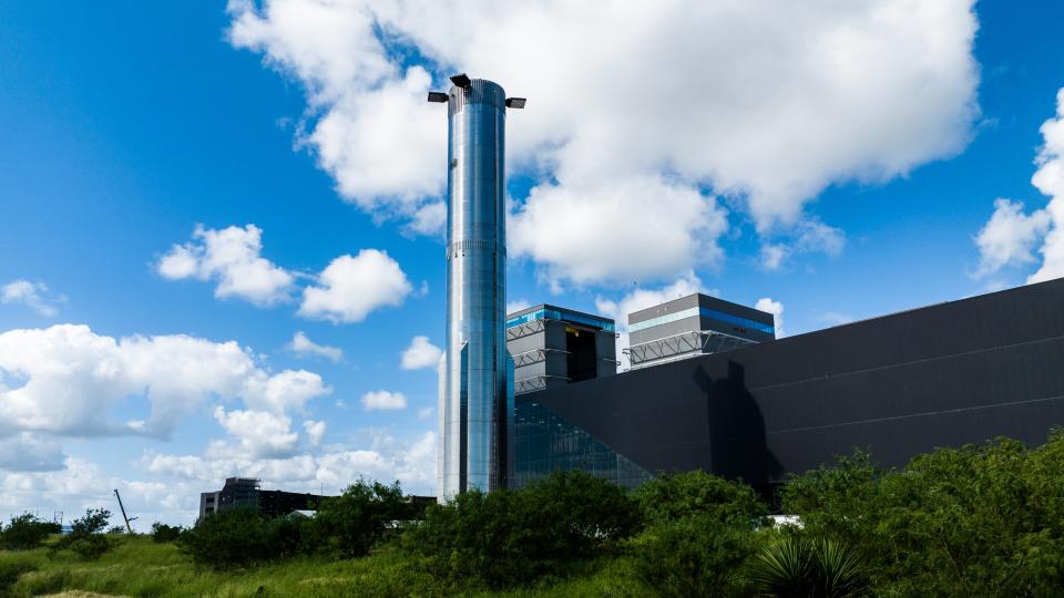  A tall silver rocket in front of a long low building and blue sky. 