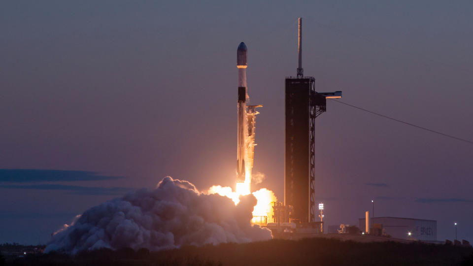  A black and white rocket launches into a darkening evening sky. 