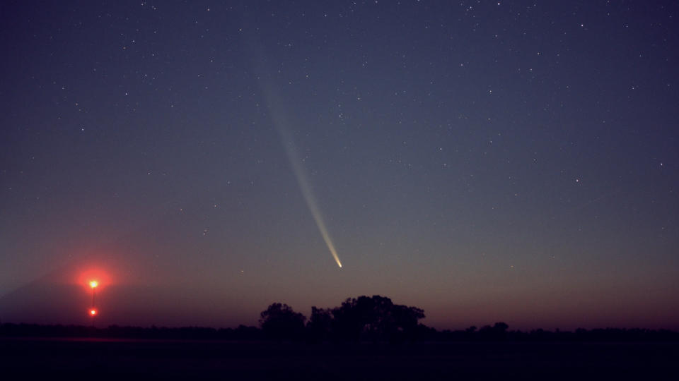  A comet streaks through a dim night sky as a faint sun sets. 