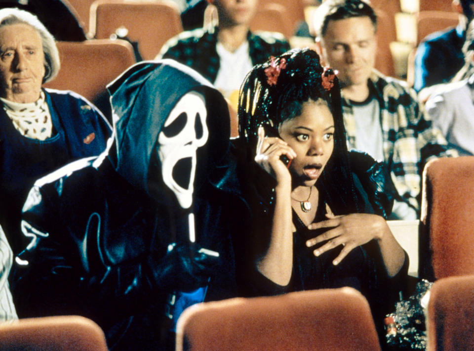 Regina Hall, looking horrified, sits next to a hooded skeleton figure in a movie theater. 