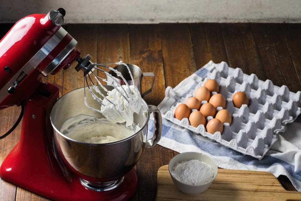 Standing mixer with cream on whisk on wooden table with ingredients.