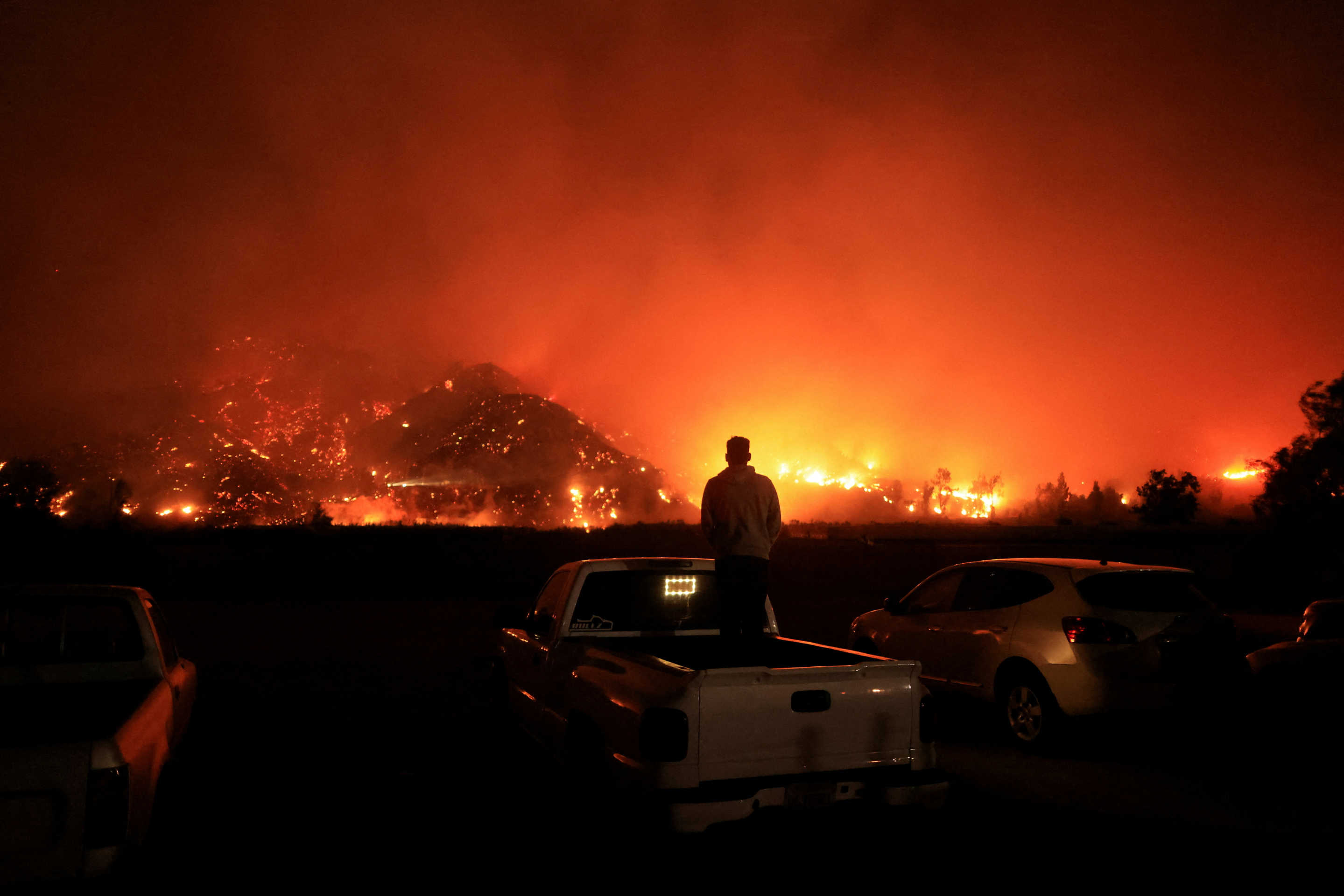 A person standing in the back of their pickup truck looks on as smoke and flames billow from the Mountain Fire in the distance.