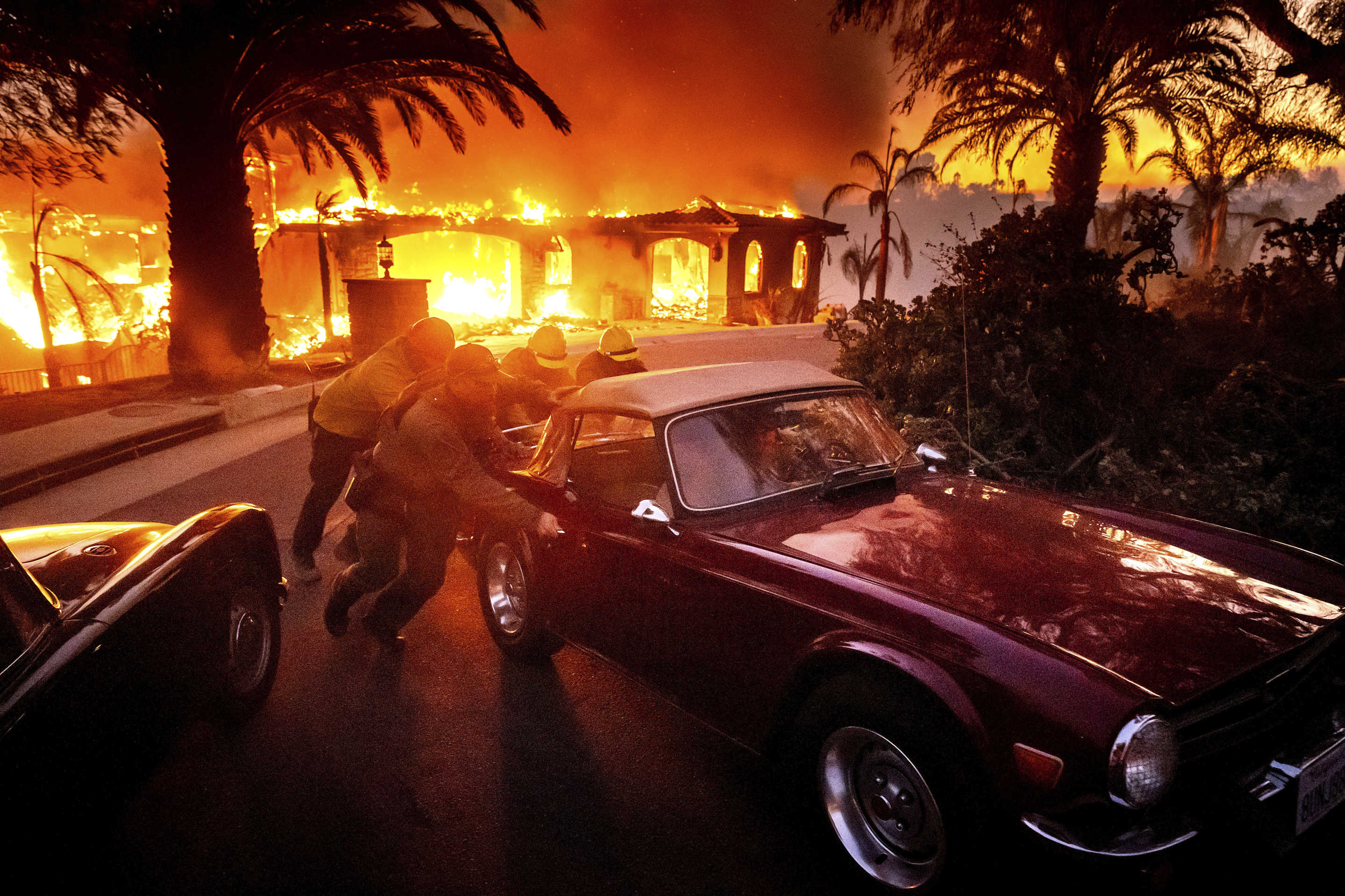 Firefighters and sheriff's deputies push a vintage car away from a burning home.