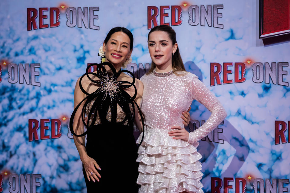 Lucy Liu, in a sleeveless evening dress with an extravagant black bow covering the top half, and Kiernan Shipka, in a sparkly pink long dress with a skirt of frills, pose on the red carpet in front of a wall showing a flocked fir tree.