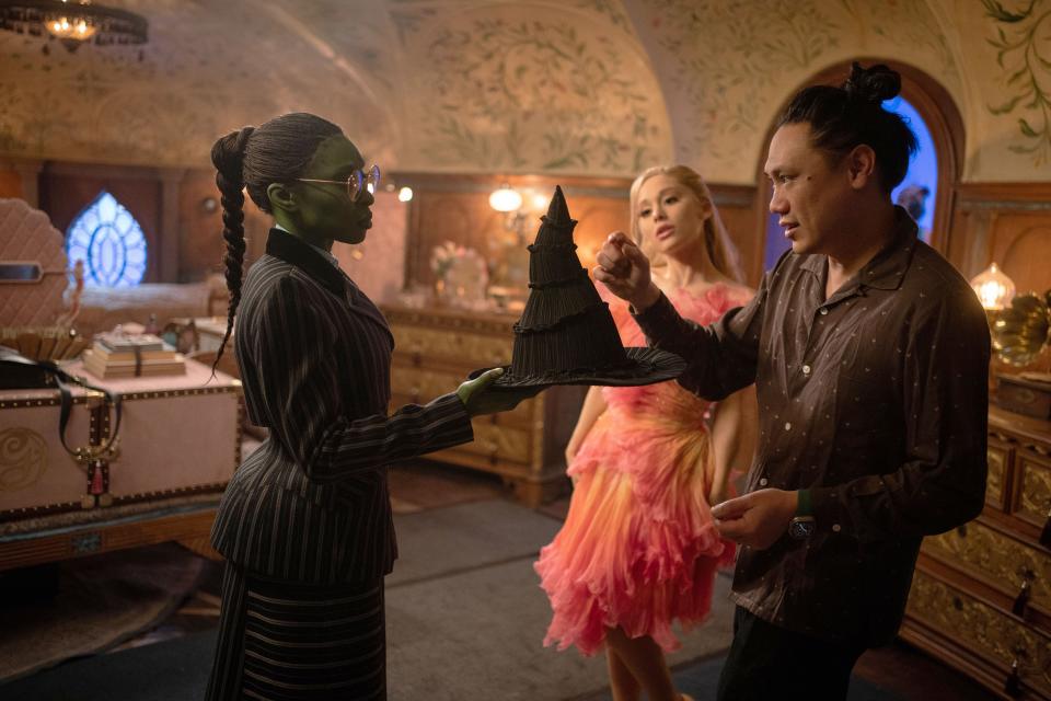 Jon M. Chu talks as Erivo, dressed in a long, fitted suit, holds out her witch's hat to him, and Grande, in pink tulle, looks on. (Giles Keyte/Universal Pictures/Courtesy Everett Collection) 