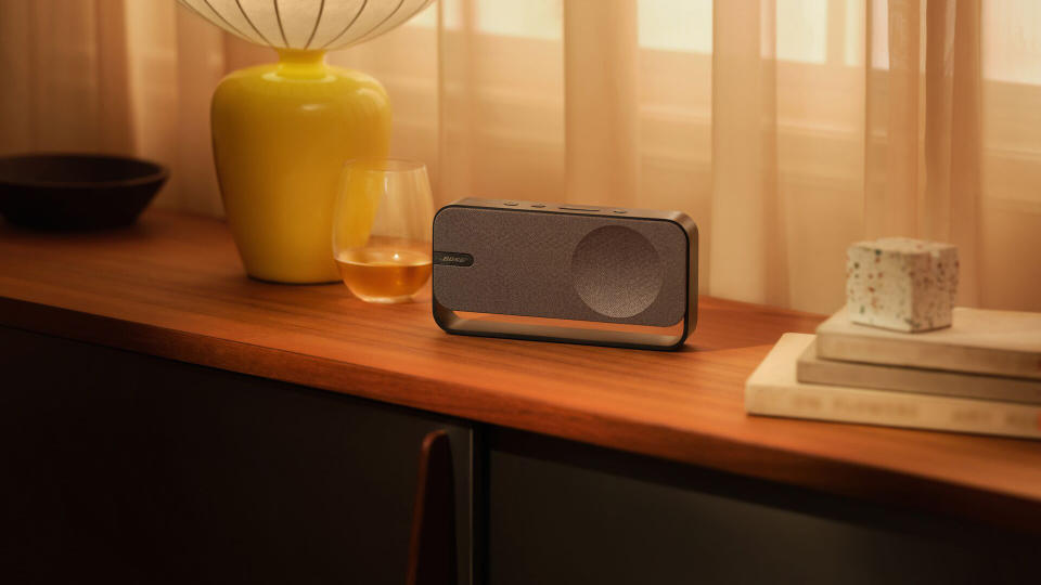 The Bose SoundLink Home speaker sitting on a shelf next to a glass. Lifestyle marketing image.