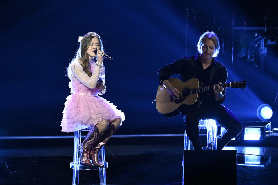 Sydney Sterlace performs sitting on a clear plastic stool in a pink frilly dress, with a guitarist beside her.