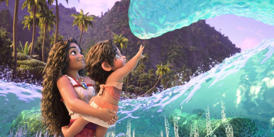 Moana, standing in the ocean, holds the baby Simea as she reaches up toward a huge breaker. 