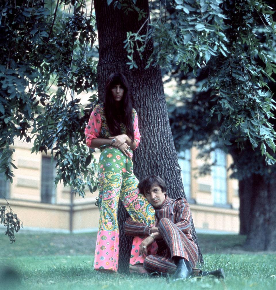 An old photo of Sonny & Cher, from the 1960s or 1970s, has Cher standing in front of a tree, with Sonny Bono sitting on the ground with one arm wrapped around Cher's leg.