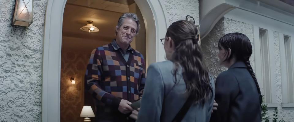 Hugh Grant in checkered cardigan, opens his front door obligingly to Chloe East and Sophie Thatcher.