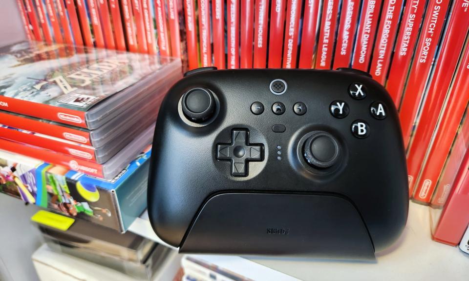 A black video game controller, the 8BitDo Ultimate Bluetooth Controller, rests on a white shelf in front of a row of several red Nintendo Switch video game cases.