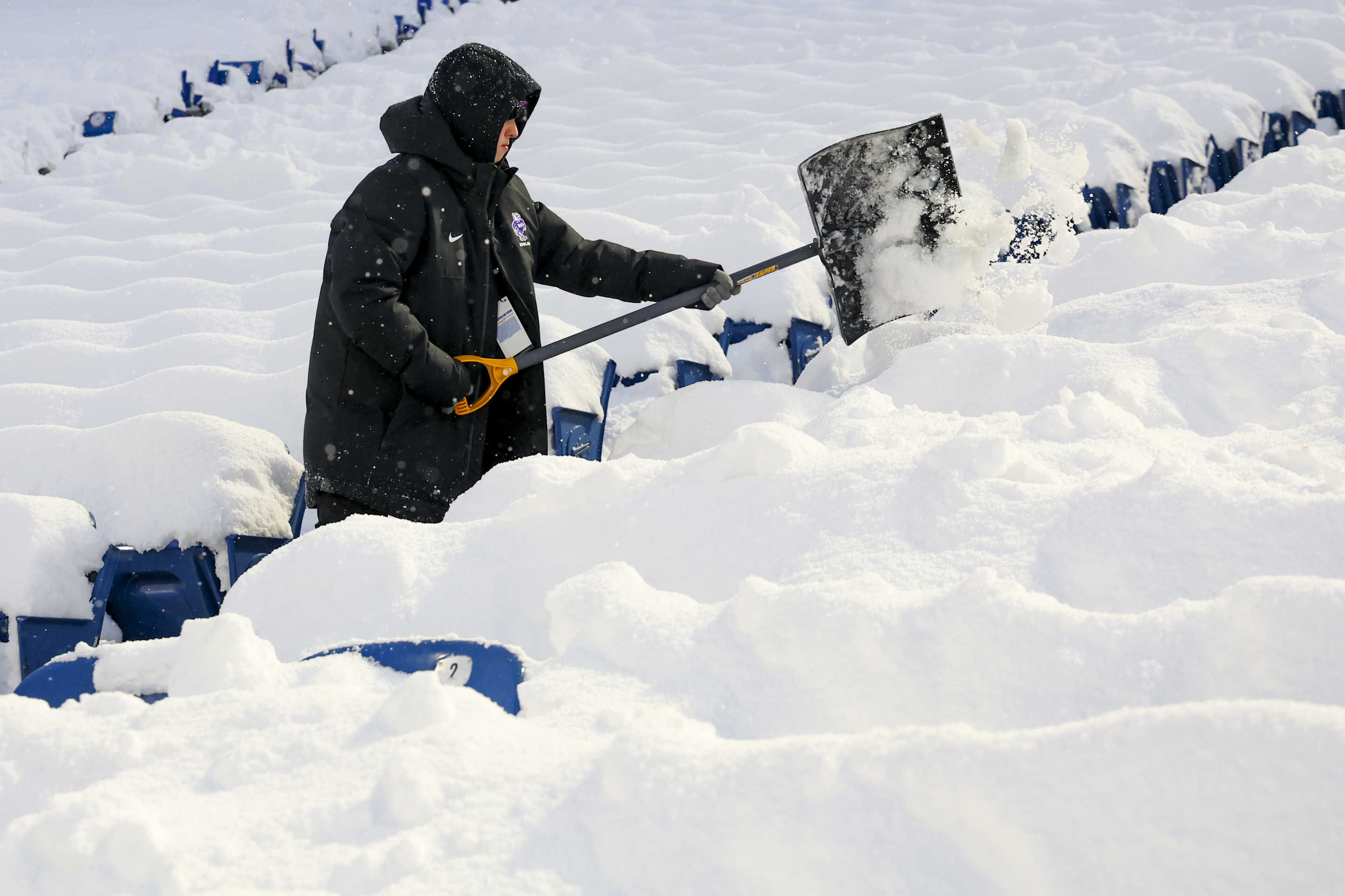 A person bundled up with a jacket, hat and gloves tries to clear the snow at Highmark Stadium with a shovel.