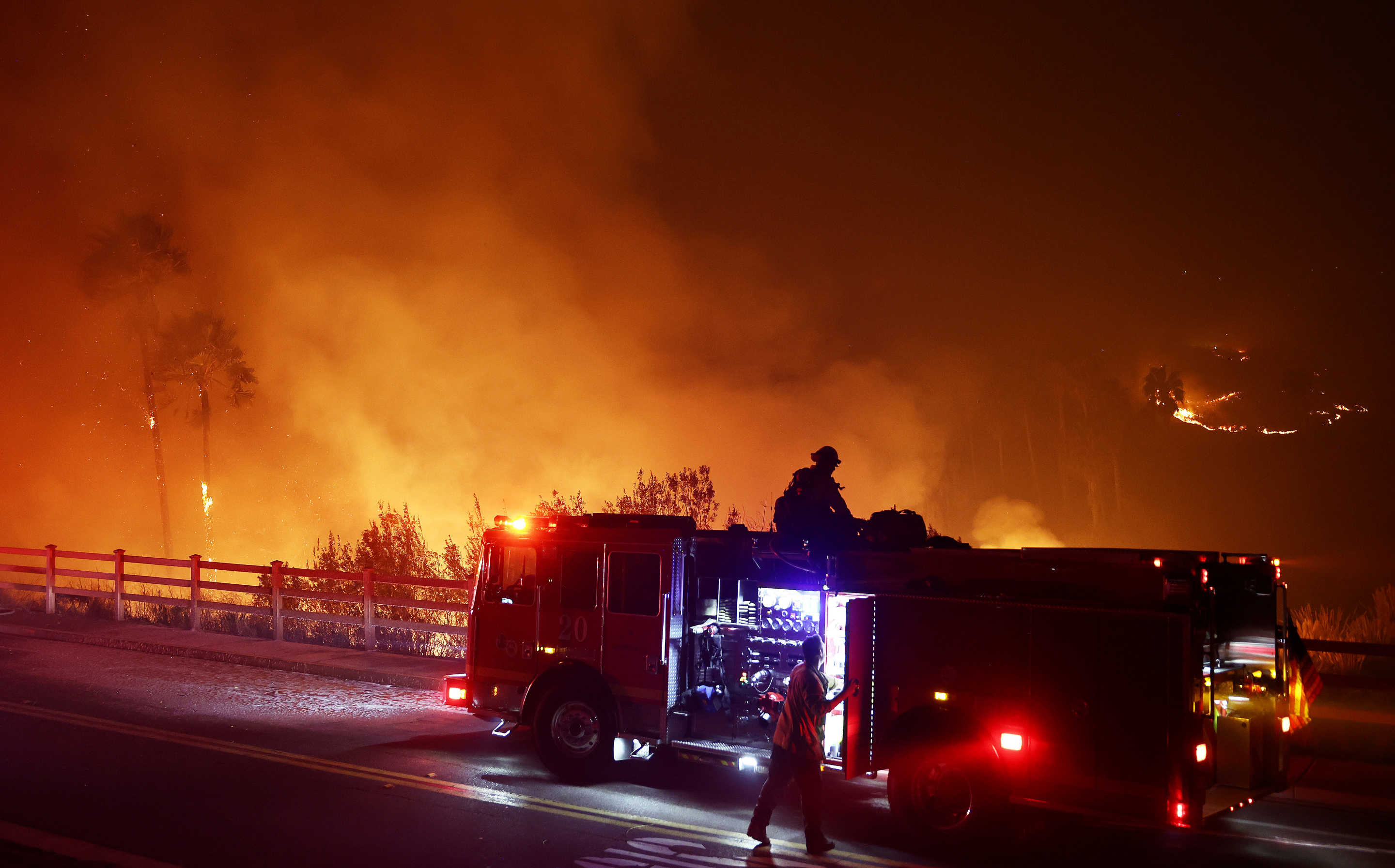 A firefighter is silhouetted on the roof of a fire truck, with smoke rising behind.