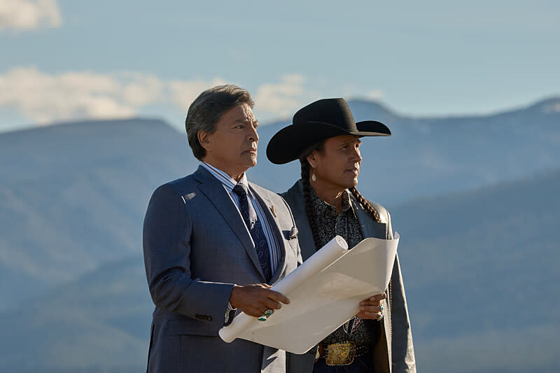 Against a backdrop of mountains, Gil Birmingham, in suit and tie, holds a scroll of paper and what looks like a title deed, as Mo Brings Plenty, in cowboy hat and braids, looks into the distance.