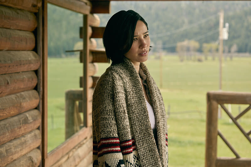 Kelsey Asbille, wearing a knit duster with Native American motifs, looks wistfully out from the porch of a log cabin.