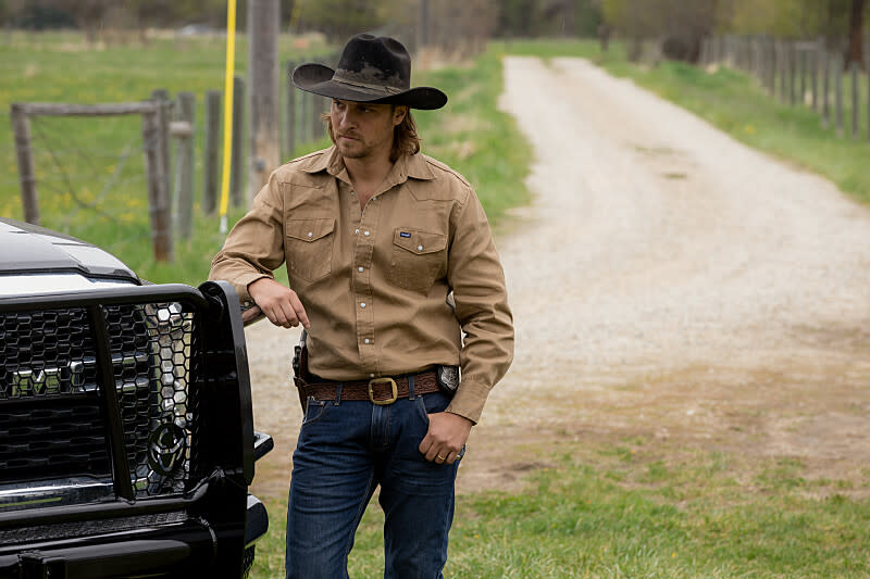 Wearing his ranger's hat and jeans, Luke Grimes leans against his vehicle on a dirt road with fields separated off with wood fences.