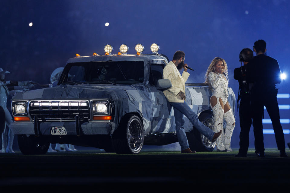 Post Malone, in jeans, leans back against a pickup truck topped with searchlights, next to Beyoncé, who is wearing white sequinned pants with cutouts at the top of the thighs.a