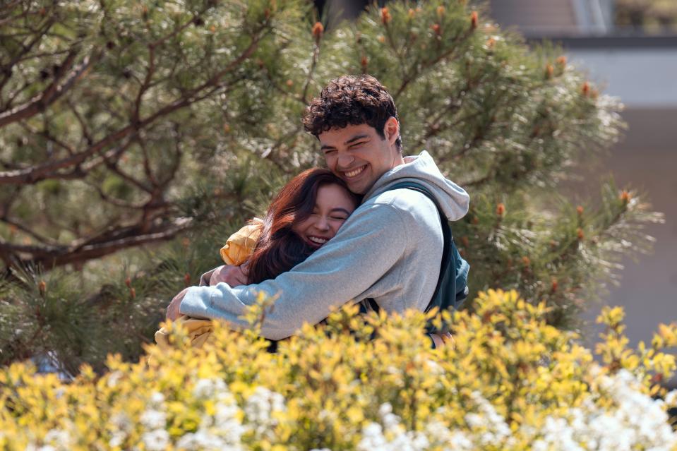 Noah Centineo, in blur hoodie and backpack, hugs Anna Cathcart in front of a yellow shrub with an evergreen tree in the background.