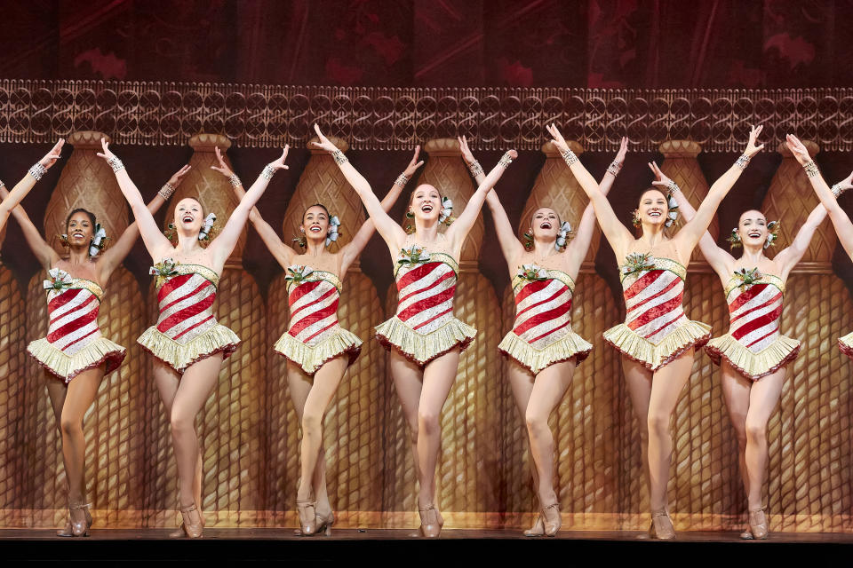 A half dozen Rockettes onstage raise their arms wearing holiday attire.