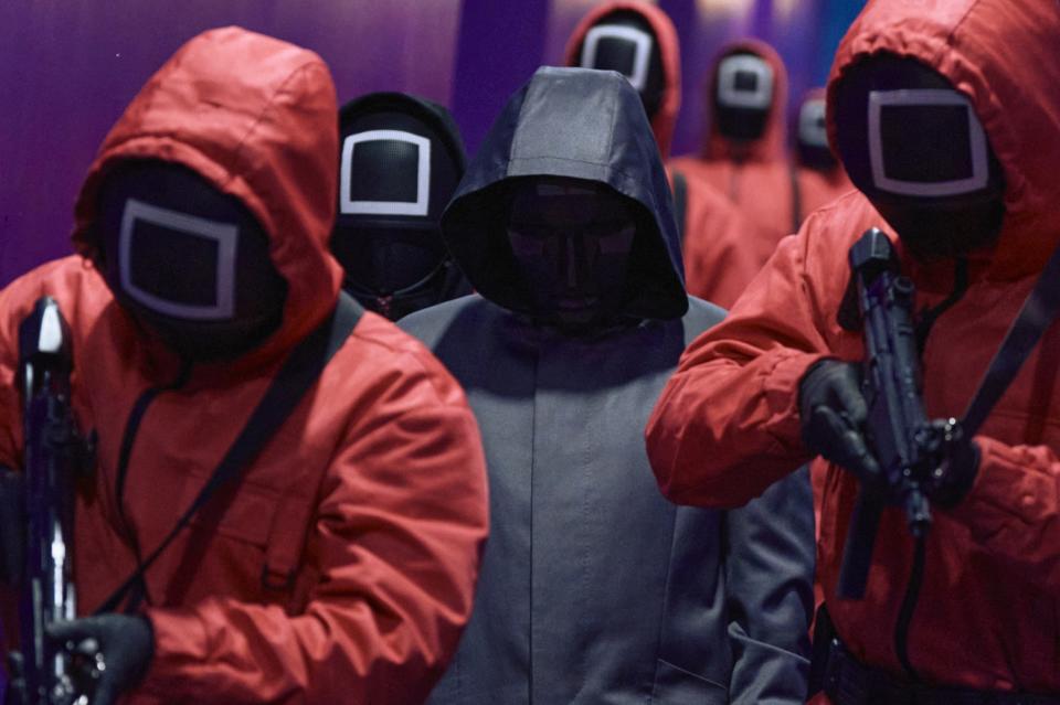 A half-dozen people in hazmat suits with hoods and masks, with one in the center wearing gray.