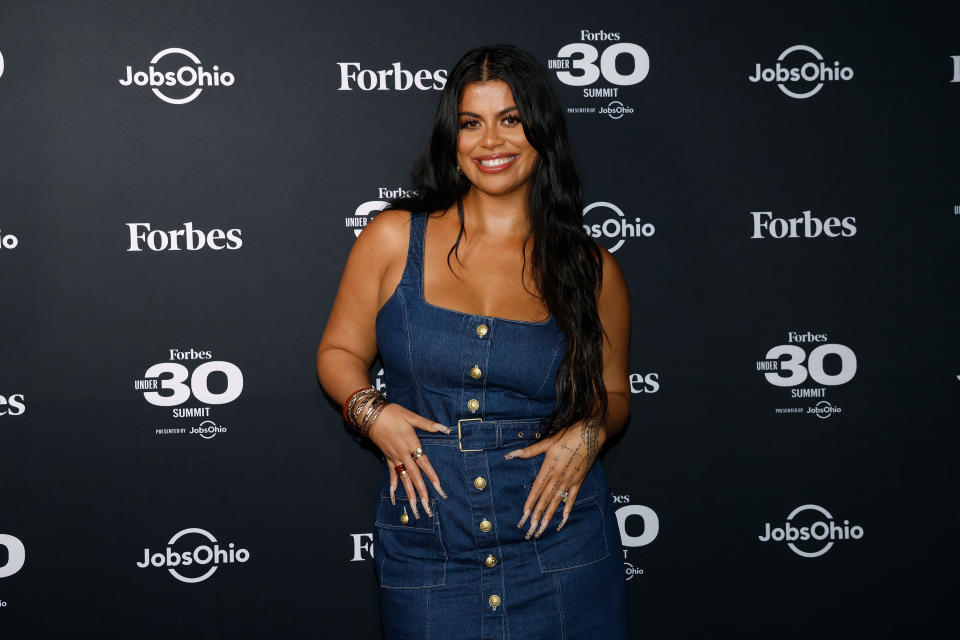 Drew Afualo, in a spaghetti strap denim dress, metal bracelets and extra-long fingernails, poses on a Forbes Summit red carpet.