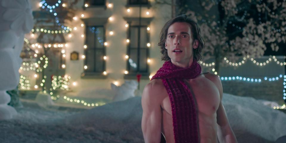 Dustin Milligan, shirtless with wool scarf, outside in the snow with strings of Christmas lights on the building in the background.