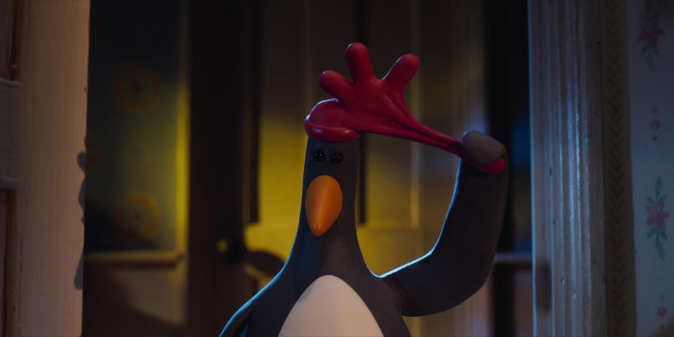 A stop-animation penguin holds the beak of a red rubber cock's crest placed on his head.