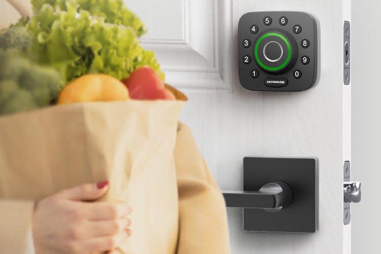 A person holding a bag of groceries stands in front of an open white door with an Ultraloq keypad deadbolt installed on it.